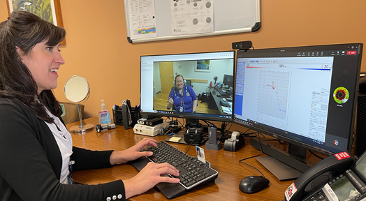 White female provider with brown hair sits in front of computer monitors and preforms video visit 