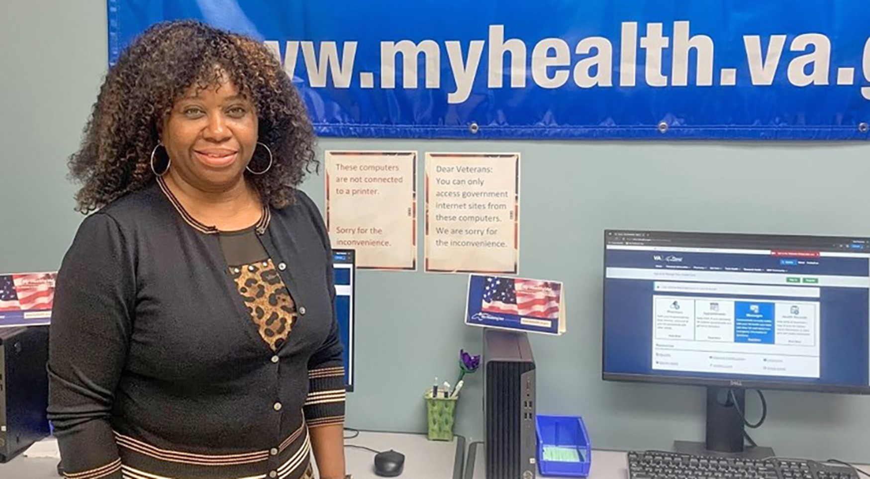 Middle aged Black woman stands in front of My HealtheVet banner in a VA facility