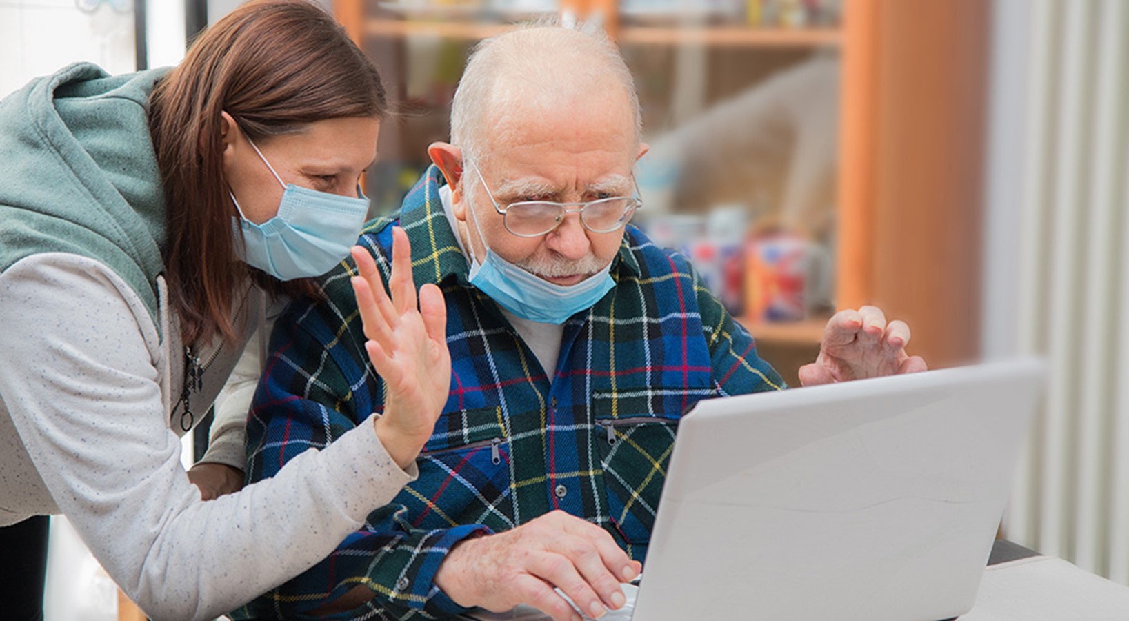elderly man sits at computer while younger woman leans over his shoulder.
