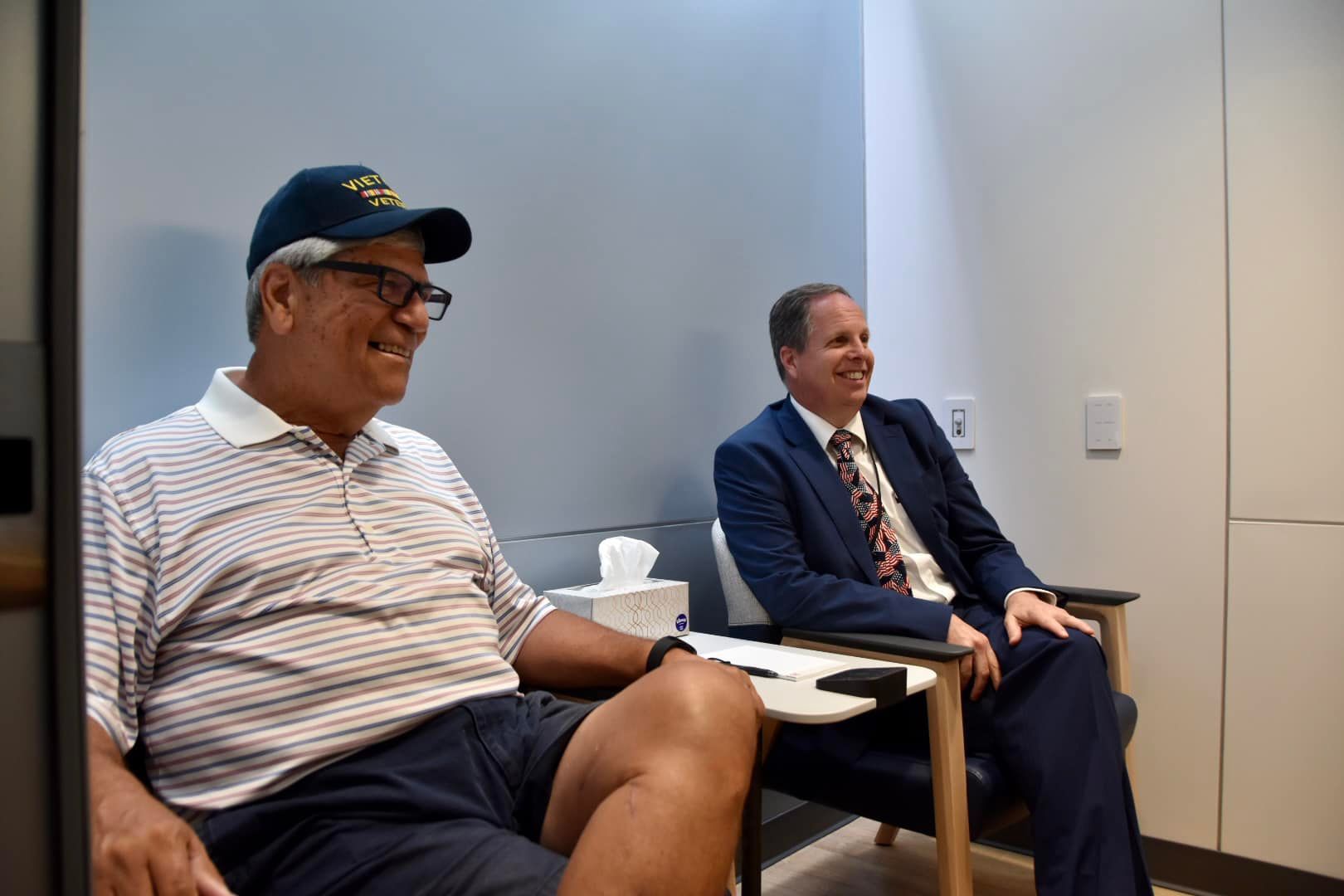 Middle aged men, one in casual clothing and one in a suit, sit inside a room next to each other 