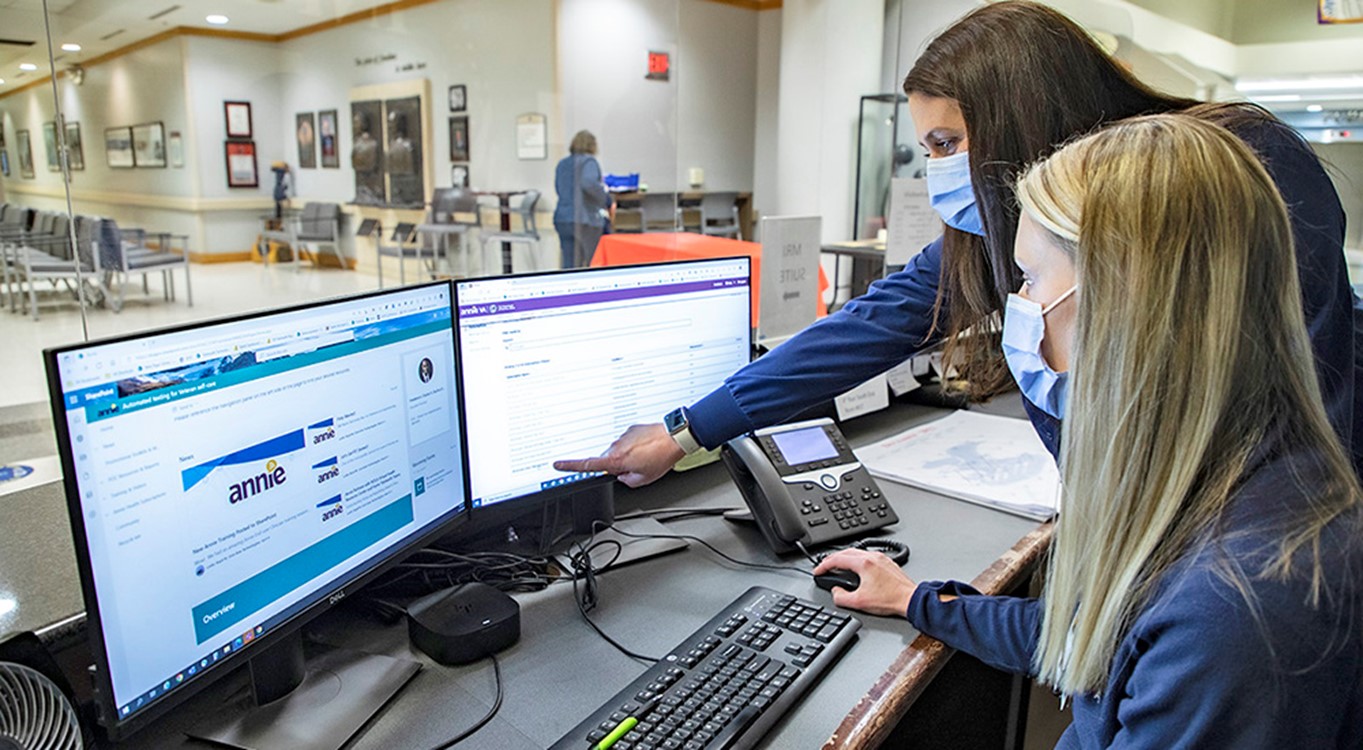 One nurse sits in front of a computer screen, while another nurse points to the screen