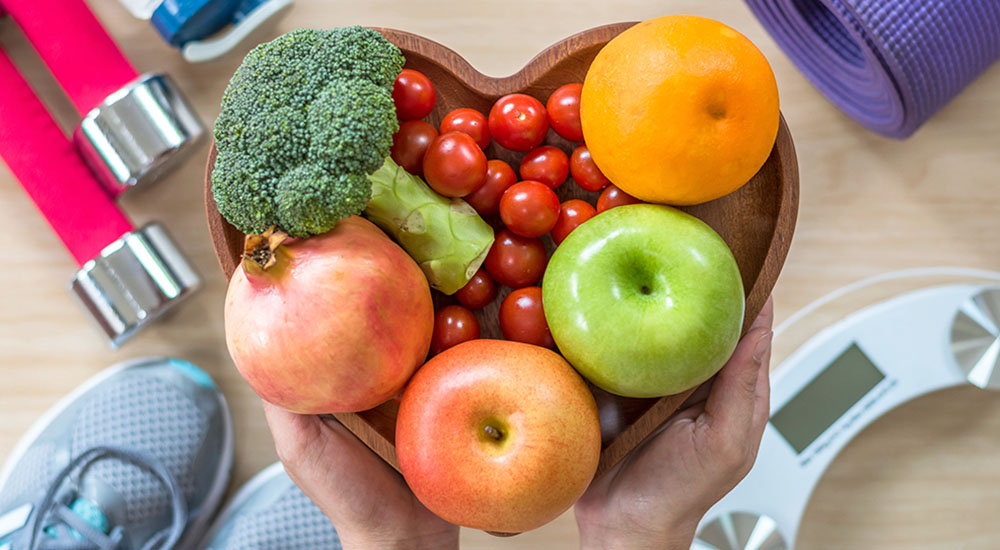 Heart-shaped bowl holding apples, an orange, tomatoes and broccoli
