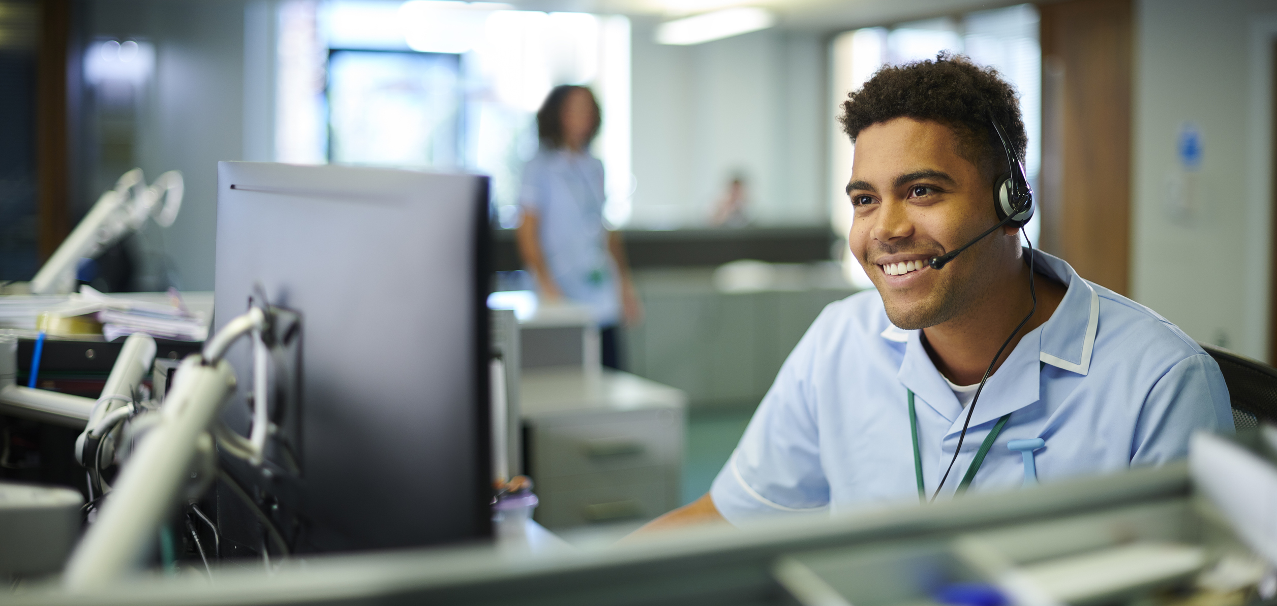 young Black man sits in front of desk with headset on, smiling