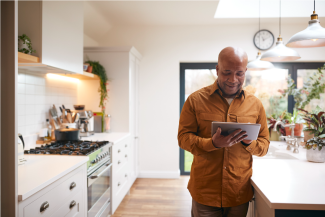 Middle aged Black man with orange shirt holds and looks at tablet in kitchen 