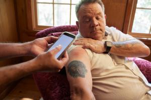 Older man sitting in a chair rolls up his sleeve for caregiver to take picture of arm