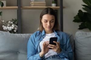 Younger woman with short, dark brown hair sits on couch and looks at cellphone