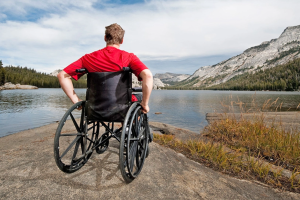 man in bright red shirt in wheelchair in front of body of water and mountains 