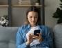 Younger woman with short, dark brown hair sits on couch and looks at cellphone