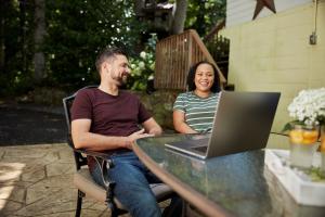 man and woman sit outside and smile at computer on a table in front of them