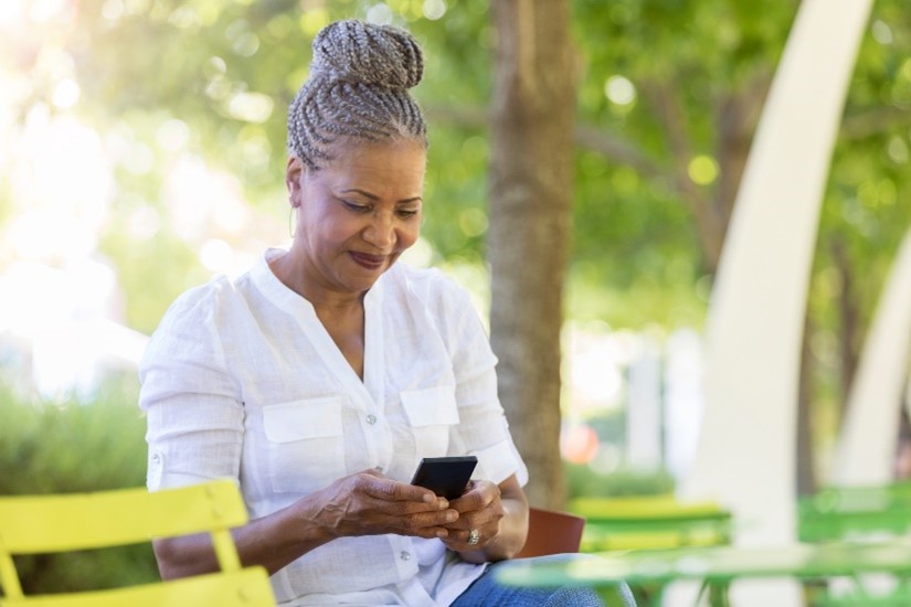 Middle-aged Black woman sites on bench in a park and smiles at her phone 